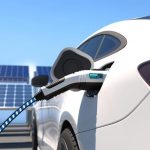 Valkoinen sähköauto latauksessa aurinkopaneelien edessä aurinkoisena päivänä.