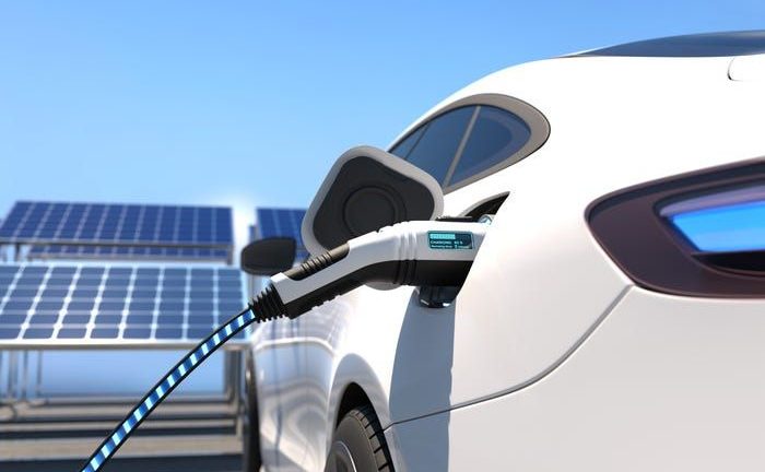 Valkoinen sähköauto latauksessa aurinkopaneelien edessä aurinkoisena päivänä.